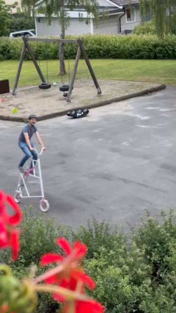 Man cycles in a circle around a kids playground on a tallbike with tiny wheels and very low gearing, resulting in rather a lot of pedalling for very little movement. 