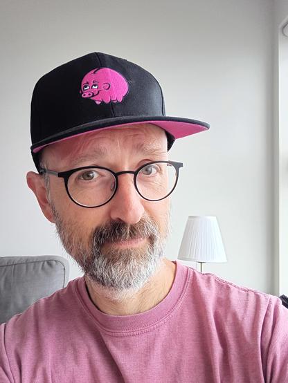 Man with glasses looking at the camera wearing a black cap with pig logo. 