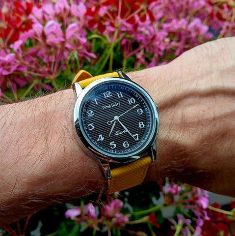 Widdershins running watch with a black face, silver case and yellow strap, on a hairy arm. Time shown os 4:33 [PM]. In the background are pink and red flowers. 