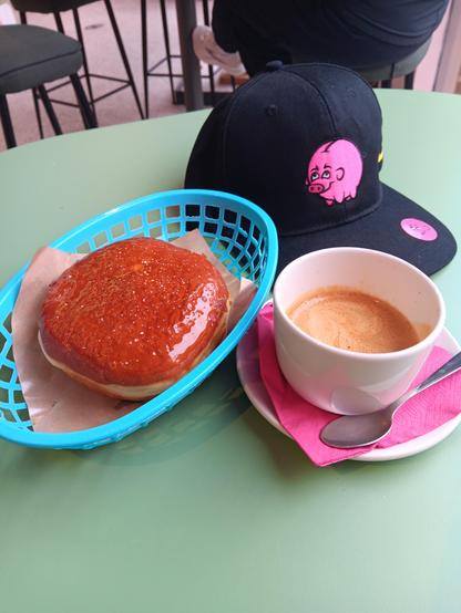 An image of A coffee and a donut next to a black baseball cap with a pig on it. 