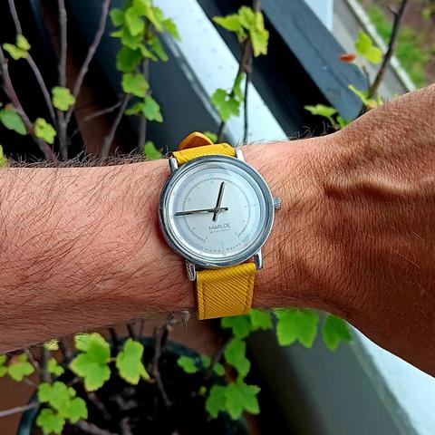 Gray dial watch with reflective steel hands and minimal guage like markings with a bright yellow leather strap, on a slightly hairy wrist. There is a small green plant in the background. 