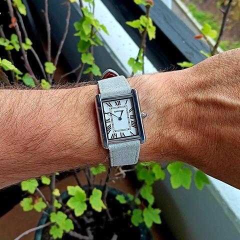 Sanda branded watch that clones the classic Cartier "Tank" styling, being small and rectangular, with Roman numerals, inner minute track, plus short blue hour and minutes hands (no seconds) and a polished metal case. The strap is light gray and has a suede like appearance. It is worn on a slightly hairy wrist. There is a small green plant in the background. 