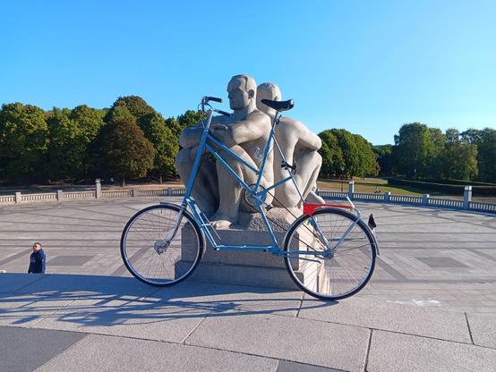 A blue framed tallbike, resting on a stone statue of a man sitting and resting his hands on his knees, from Vigelandsparken. It is a sunny day with clear skies and there are many trees in the background