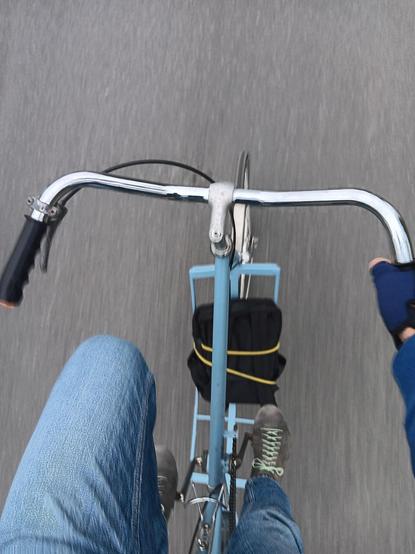 Looking down at a blue tall bike from the point of view of the rider. It is possible to see the frame of the bike and one hand on the handlebars, along with the riders legs. Below that is a small black backpack strapped to the bottom of the frame.