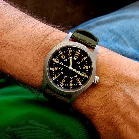 A field watch with a black dial and numerals for 12 and 24 hour. Along with numbered minutes at every 5 intervals. On a green rubber strap. On a fairly hairy wrist. In the background you can make out the wearer has a green T-shirt and blue jeans. 