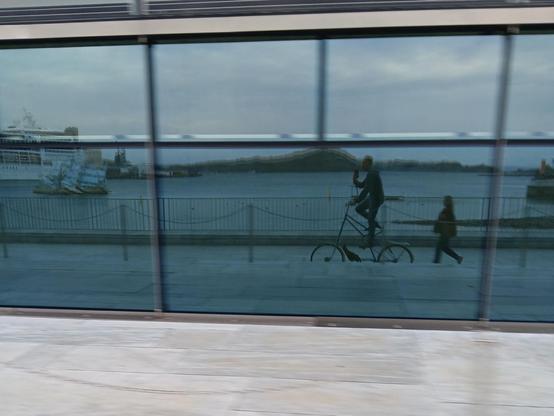 Reflection on a large glass wall showing a waterfront scene with a person riding a tall bicycle and another walking beside them. In the background, the stainless steel and glass sculpture "She Lies" by Monica Bonvicini floats in front of a docked cruise ship, with a distant hill visible across the water.