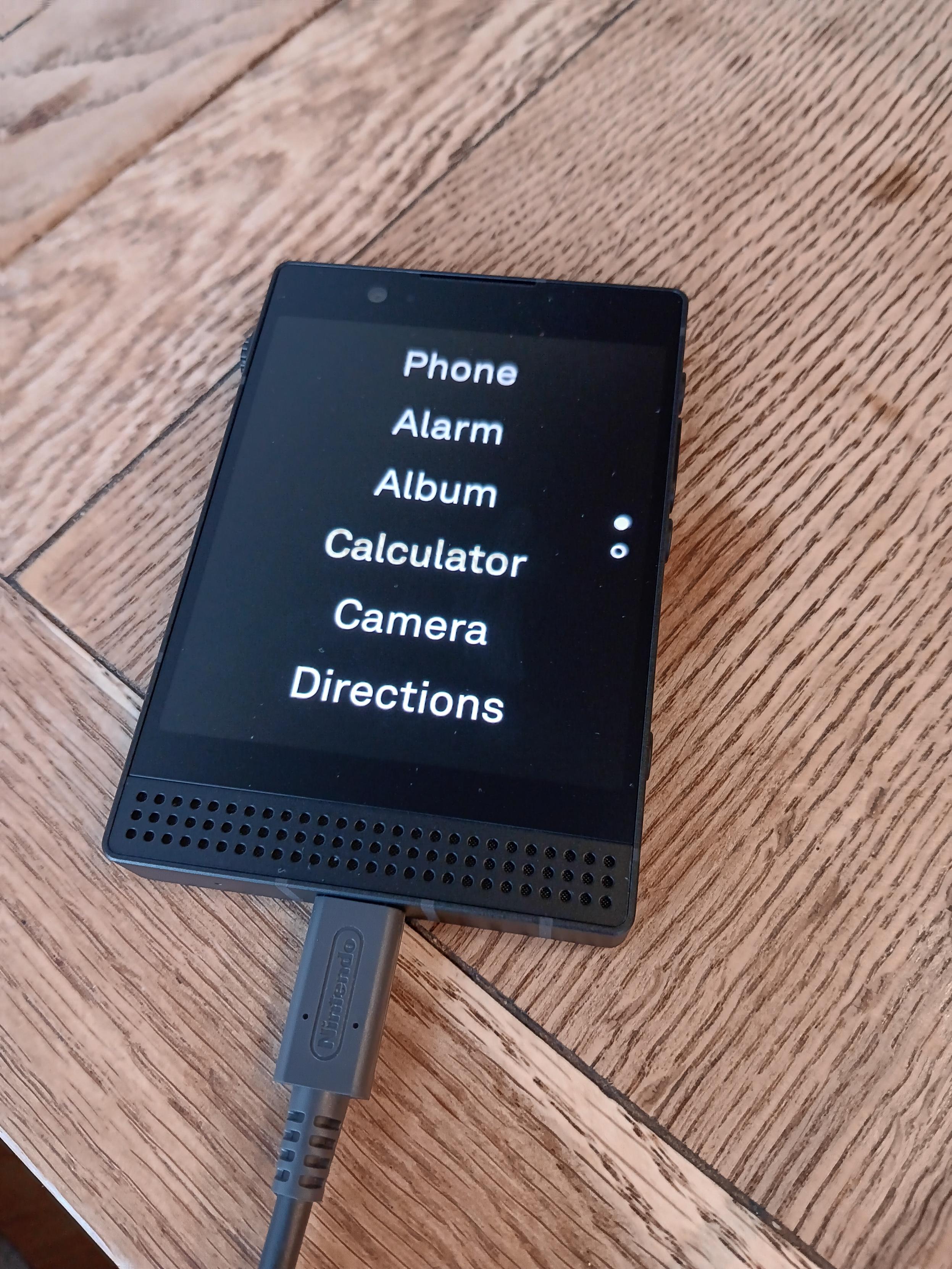 Lightphone III on a wooden desk showing a menu of phone, alarm, album, calculator, camera and directions.