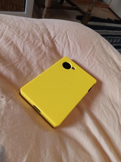 A lightphone III with a bright yellow case resting upside down. The holes for the camera and square shape give it a block of cheese vibe. Resting on a duvet cover. 