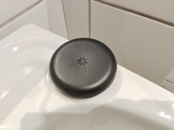 A circular, black, plastic container sits on top of a white porcelin sink. At the center of the box is a small logo that features a stylized flower-like icon with eight pointed elliptical shapes (the petals) arranged in a circular pattern. In the background white ceramic tiles can also be seen.