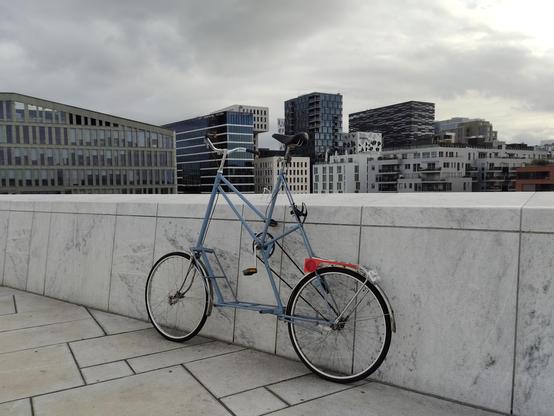 A blue tallbike leaning against white stone wall over looing some tall buildings within a city. Cloudy skies.