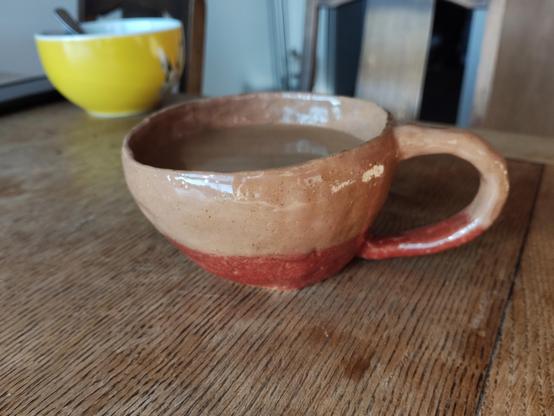 A slightly wonky looking coffee cup in the style of Quentin Blake's drawings. The lower part is a reddish brown and the upper part light in colour. Sitting on a wooden table. 
