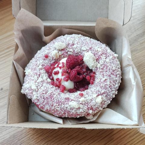 A donut with pink frosting and covered in white, sweet crumble. In the middle is vanilla cream and raspberries.