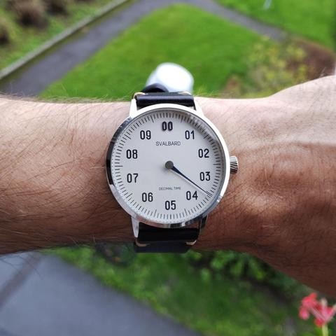 An analoge face decimal watch with a single hand showing the time as 3:56 mounted on wrist. In the background you can make out grass. 