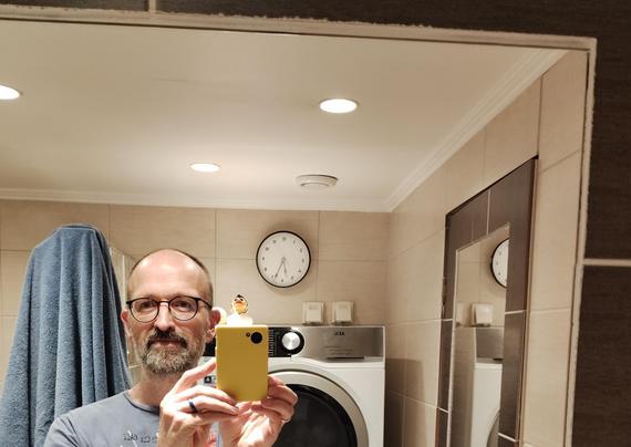 A picture taken selfie mirror style in a bathroom. In the foreground a man holding a yellow phone up. In the background a clck that shows the time as 5:34. The numbers are mirrored but the time looks as expected.