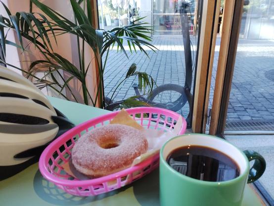 A cafe table with a pink basket holding a sugar-coated donut on paper, beside a green ceramic mug filled with black coffee. A white and black bicycle helmet rests nearby. Through the window in the background, a Halfbike is parked on cobblestone pavement, partially obscured by green plants.