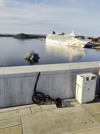 Scenic waterfront view from the rooftop of the Oslo Opera House. In the foreground, a Halfbike leans against a white stone wall, next to a bin. In the  water below is “She Lies” (a glass and steel sculpture resembling a fragmented iceberg). In the background, a large cruise ship is docked at the port, with hills and city buildings visible under clear daylight.
