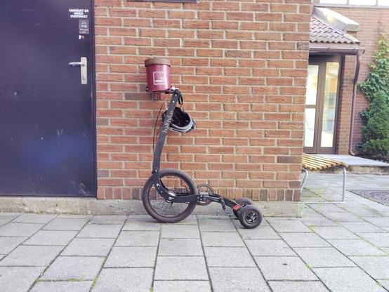 A Halfbike leans against a brick wall on a tiled pavement. A red cylindrical ciggerette bin is is just in front of the bike frame, and a black helmet hangs from the handlebar. The setting includes a black door to the left and a building with large windows and a bench to the right.