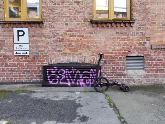 A black Halfbike (a type of stand up tricycle) in front of an old brick building. It is next to some black painted ventilation point for the building, that is covered in indecipherable pink graffiti.