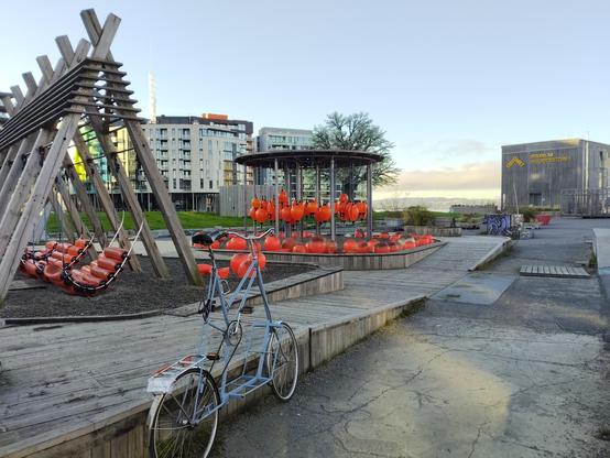 A pastel blue tallbike parked next to a wooden platform in an urban play area built in part with red marine floats. Modern buildings frame the background. The skies are clear and lighting is such that you can tell this is from early morning.