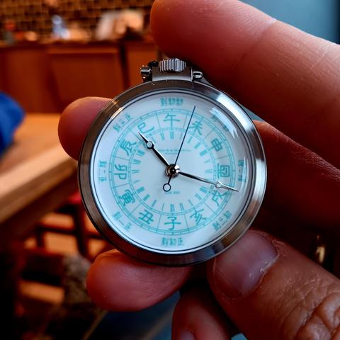A pocket watch held in hand, behind which is an unfocussed room. The watch has twelve Shíchen (double hour) markers represented by the Earthly Branches, as also used in the "Chinese zodiac". Inside those markers it has two sets of twelve Roman Numerals to show the twenty-four "normal" hours of a day [the watch has a twenty-four hour movement, so the hour hand only completes one revolution per day]. Around the very outer edge are eight Kè references to represent the total per Shíchen and so impl…