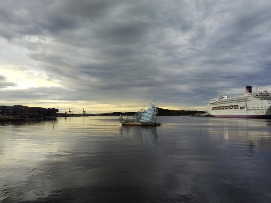 Floating in the Oslo fjord under a partially cloud covered morning sky, the She Lies sculpture (an abstract glass and steel structure resembling an iceberg) rests in the water. To the right, a large cruise ship is docked, its decks and windows visible. On the left, shoreline buildings line the harbor.