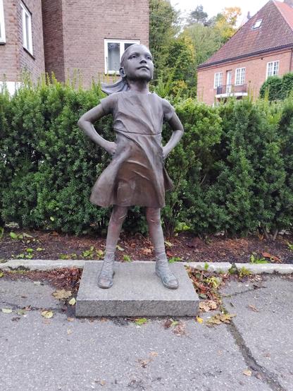 A bronze statue of a young girl stands confidently with hands on hips and feet apart, gazing upward. She is placed on a stone pedestal on a paved surface, with hedges and brick buildings behind her. 
