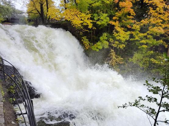 A waterfall on Akerselva (Olso's main river) thunders down a rocky incline framed by vivid fall foliage. A stone path with a metal railing curves along the left side, offering a scenic viewpoint. Mist rises from the base, emphasizing the force and energy of the water.