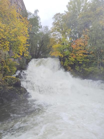 A waterfall on Akerselva cascades down a rocky slope near Beierbrua, surrounded by autumn-colored trees. White foam and mist rise from the crashing water. On the left, a partially visible old industrial building, contrasting with the vibrant natural scene.