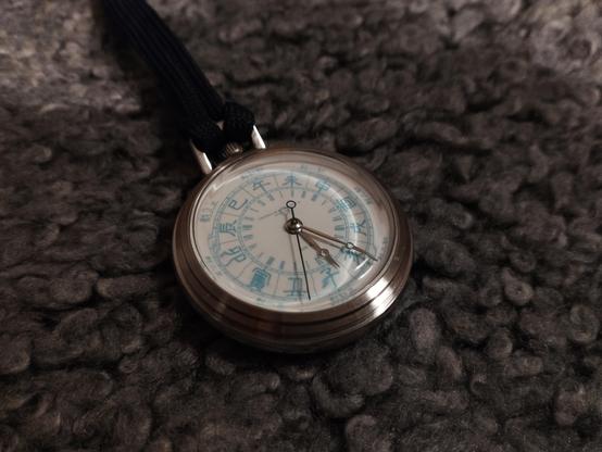 A pocket watch with Chinese Shíchen (時辰) in an outer ring and a double set of 12 Roman Numerals on an inner ring, covering a 24 hour range. The hour hand is pointing just after an Ⅺ marker and the minute hand is at 25 past. However since the hour hand is within the 子 area, we know this is night and thus 11:25PM/23:25.

The markings are all light blue on a white background. There is some distortion due to the domed crystal and the viewing angle. The watch has a steel case and is attached to a da…