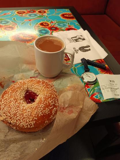 Peanut and jam filled bagel with a sesame coating and a cup of coffee, resting on brown paper. Next to them are napkins, a receipt and a pocket watch. All resting on a floral patterned table. 