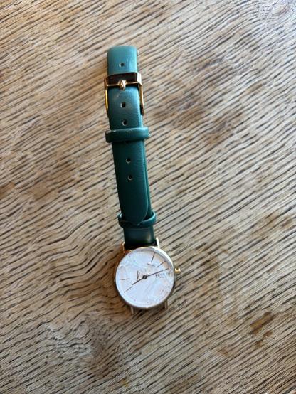 A broken watch on a wooden surface. It has a cracked dial and markers have fallen off.