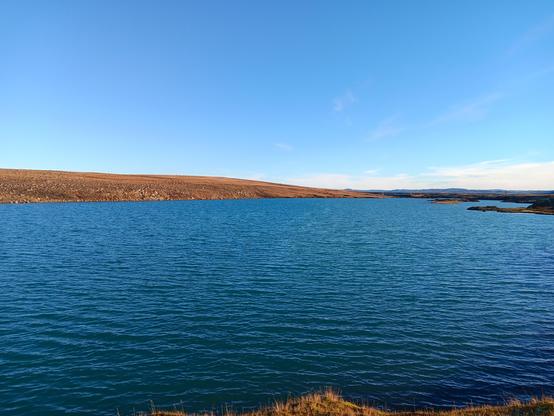 A shoreline to a body of water, with brownish, green land in the distance. The water and sky are vibrant blue.