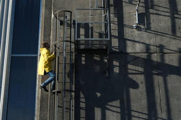 Man with yellow jacket climbing on the outside of a ladder up an industrial looking building. 