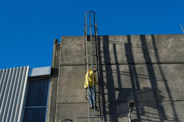Man with yellow jacket climbing up a ladder, up an industrial looking building. Very blue sky. 