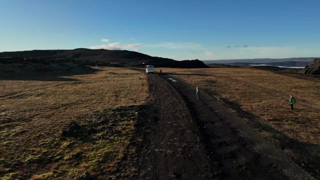 A drone follows a man on a unicycle, cycling along a dirt path up to a white bus in Icelandic nature on a sunny day. 