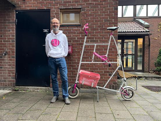 Middle aged, beared, white man looking at the camera and smiling. He is wearing a white hoodie with a pink, piggie bank logo (Bónus) and blue jeans. He is standing next to a pink and white tallbike with tiny wheels. It also has a pink basket.