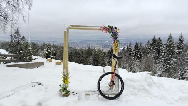 A red unicycle leaning against an empty wooden frame, decorated with flowers. Through the frame (and around) is a view looking down on Oslo. Snow on the ground and the trees.