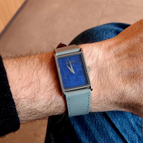 Tank style watch with a mosaic, lapis lazuli face and  gray leather strap on a hairy wrist. Jeans and a stone tiled floor visible in the background. The time on the watch is approximately 10:59. 