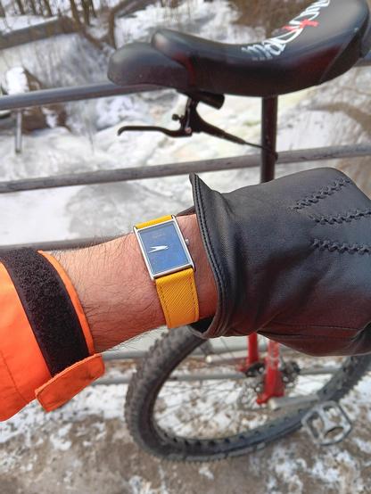 A man's wrist wearing a tank style watch with an aquamarine dial and a bright yellow strap. He is wearing a black glove and a florescent orange jacket (just visible further up his arm). In the background is a red unicycle with a studded tyre and disc brake. Behind that an icey river. 