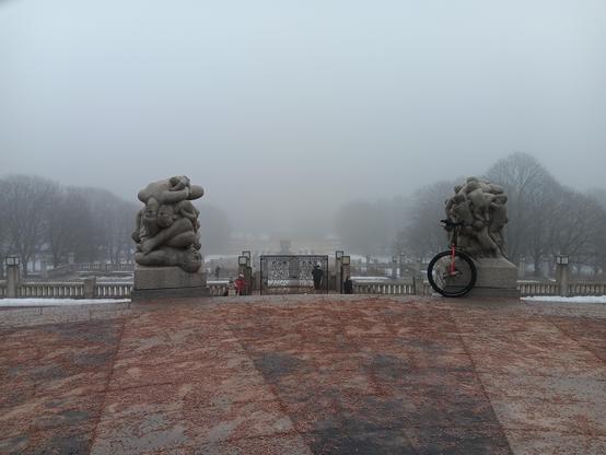 Looking out over a misty park. In the foreground a unicycle leans on a stone statue
