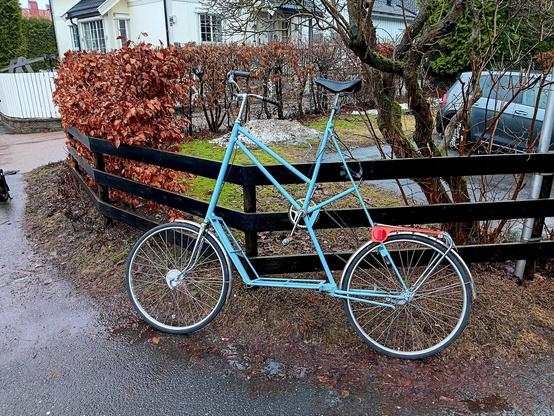 A blue tallbike leaning against a fence. There is water on the ground and the whole scene looks damp. 