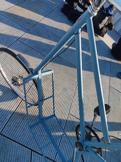 Looking down at the front of a blue, cargo, tallbike. On the ground are slatted, white stone tiles. At the edge of the picture you can see some people sitting. 