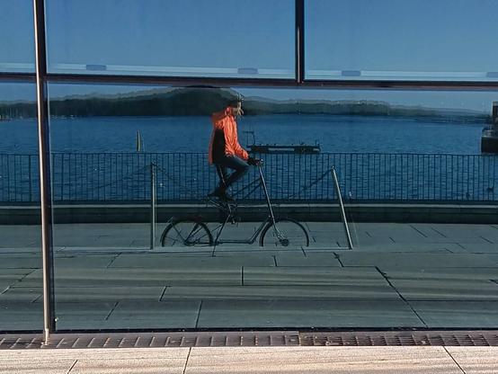 A man in a bright orange reflective jacket, riding a tallbike, taking a picture of his reflection in a large glass window. The Oslo fjord is behind.