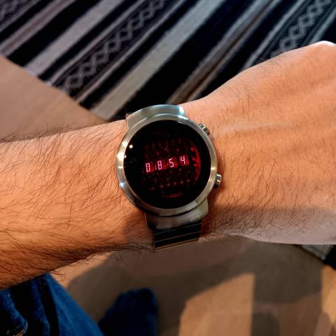 Steel watch with a circular face. The crystal is ruby red and the time is displayed with very thin LED digits (time shown is 08:54). On a reasonably hairy wrist. The picture is taken from above so that you can see the wooden floor below with a stripped rug.