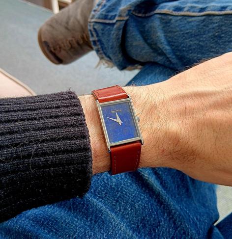 Tank style watch on wrist with a tiled, lapis lazuli dial and sword like hands. No hour or minute markers. It has a full grain 'cognac' leather strap. In the background you can see my jeans and one of my boots where my legs cross.