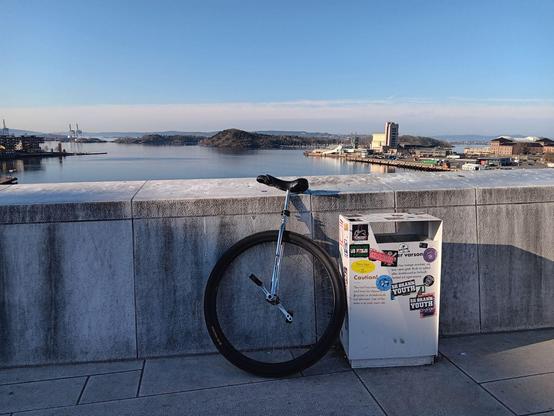 A large wheeled unicycle leaning on a white stone wall before the Oslo fjord, next to a retangular bin (trash can). The bin is covered in stickers