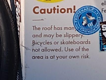 A close up of the bin from the previous image reveals a message that reads, The roof has many [hidden by a sticker] and may be slippery. Bicycles and skateboards not allowed. Use the area at your own risk.