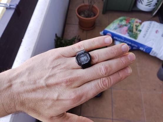 A hand wearing a ring watch that looks like a square gshock. In the background are some pots and a bag of soil.