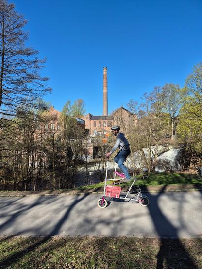 Man on a very tall bike with tiny wheels in front of a river with a waterfall. Industrial buildings are in the background.