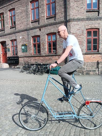 Tall bald man in a white t-shirt on a light blue tallbike with an old brick building in the background. He is cycling on road that is paved with setts.